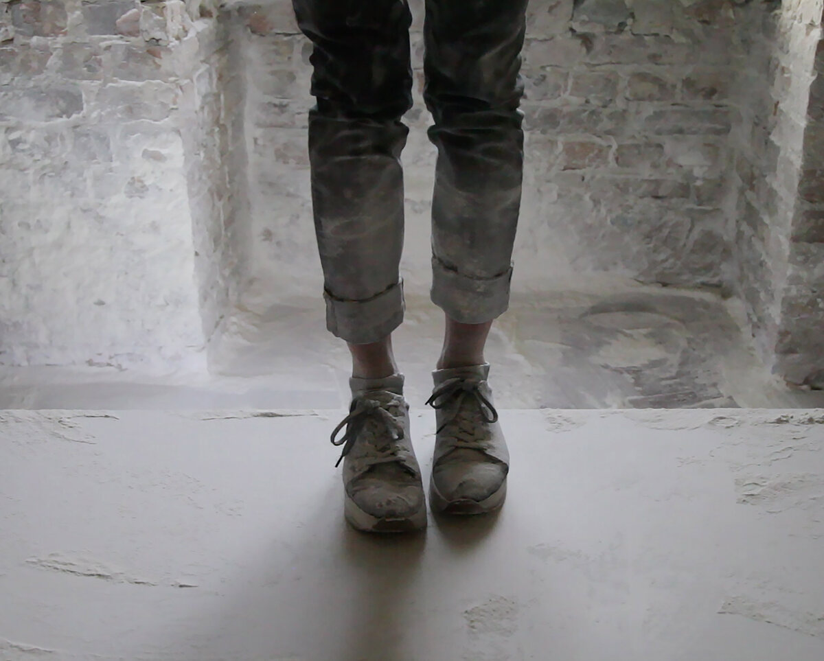 Legs in dust-covered trousers and worn trainers standing on a white plastered ledge in a bare brick room