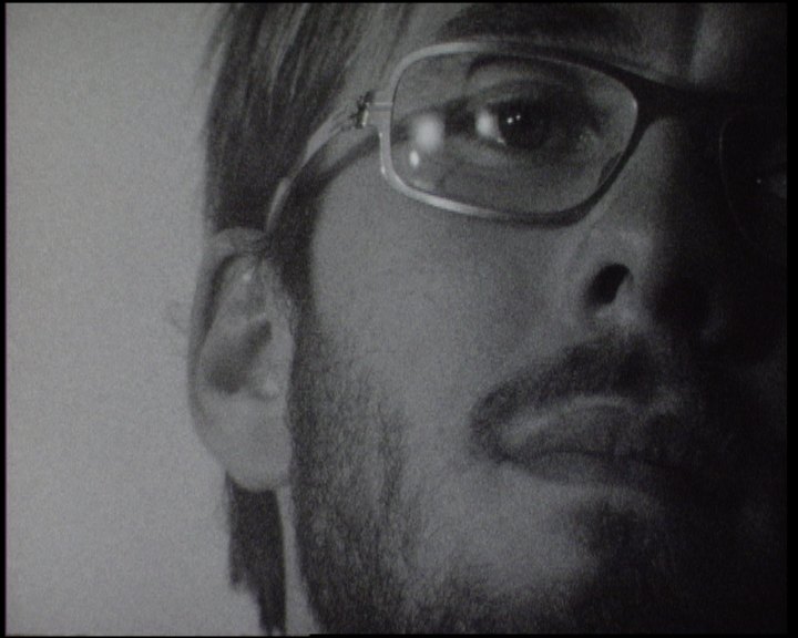 A bearded man in glasses, the frame cut just under his eyes, looking slightly past the lens