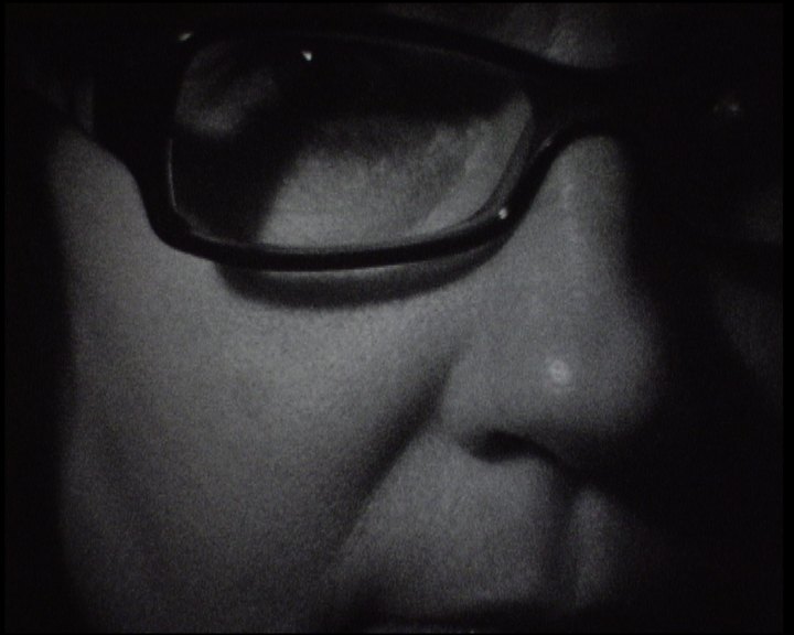 A face close to the lens in grainy black and white, dark-rimmed glasses filling the frame, a mole on the cheek, lit from the side