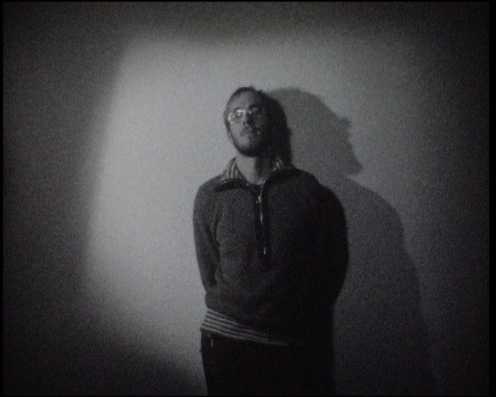 A man in glasses and a dark zip jumper standing against a wall in a circle of light, hands behind his back