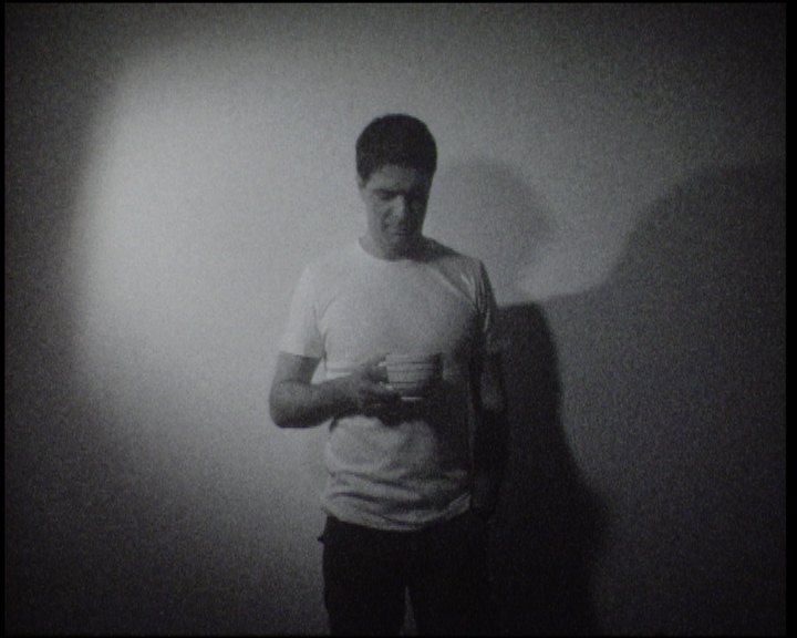 A man in a white T-shirt standing against the wall, looking down at a cup in his hands