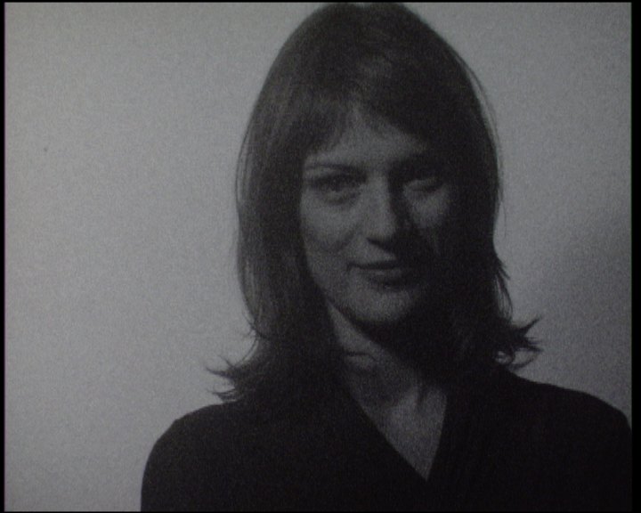 A woman with shoulder-length hair facing the camera, the wall pale behind her, a faint smile