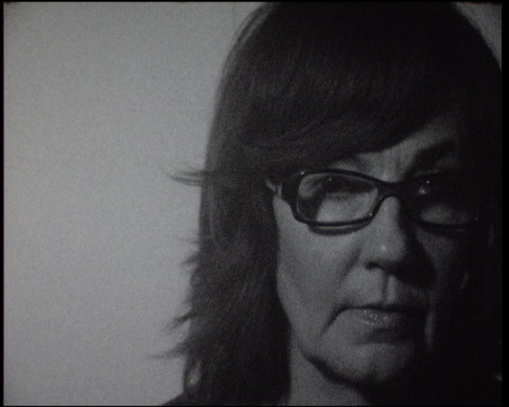 A woman in dark-rimmed glasses at the right of the frame, hair falling to her shoulders, looking straight out