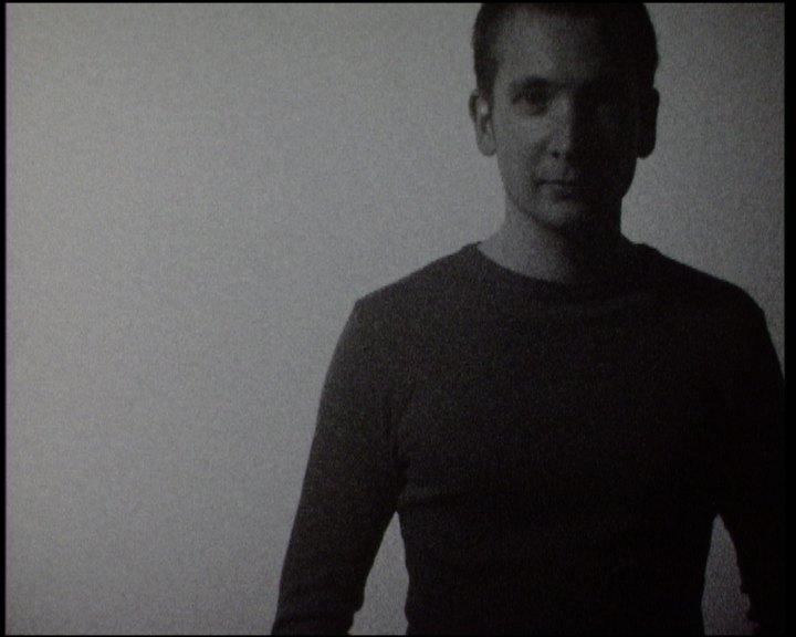 A young man in a dark long-sleeved top standing centred against the bright wall, looking into the lens