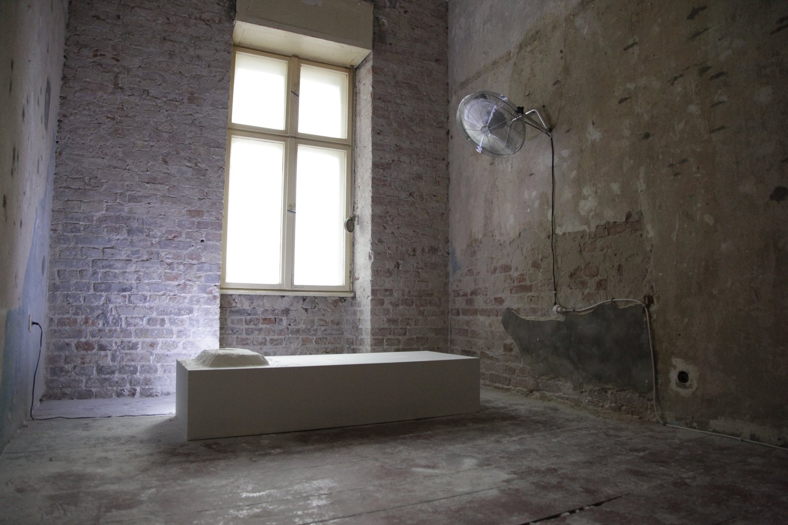 A bare room of exposed brick under a tall window: a long white platform on the floor with a heap of white powder at one end, and a fan mounted on the wall above it