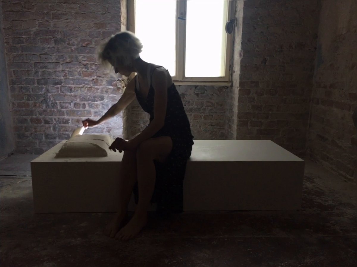 A woman seated at the end of a white platform in a dim brick room, shaping a pillow of white powder with her hands, a bright window behind her