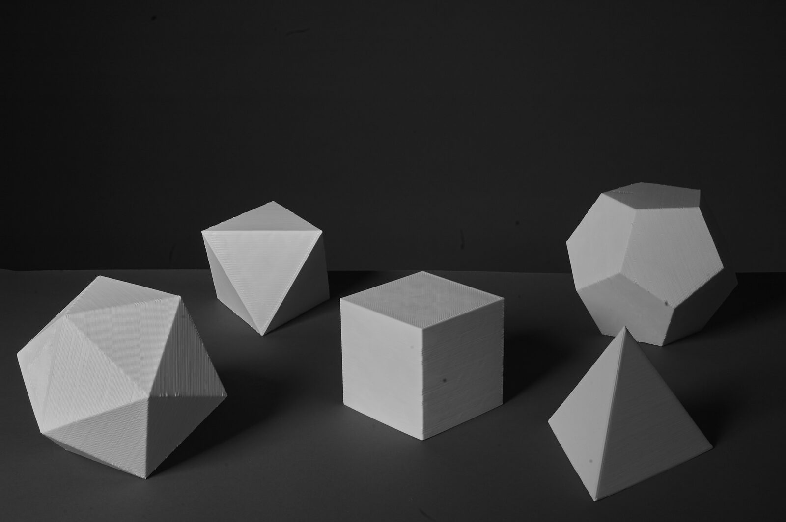 Five white 3D-printed platonic solids arranged on a grey surface against a black ground
