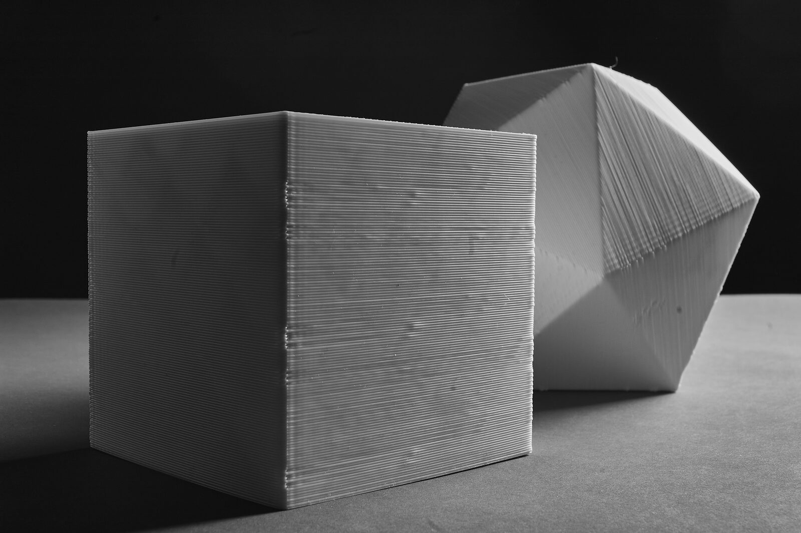 A 3D-printed cube in the foreground with an icosahedron behind it, both showing horizontal layer lines and seams