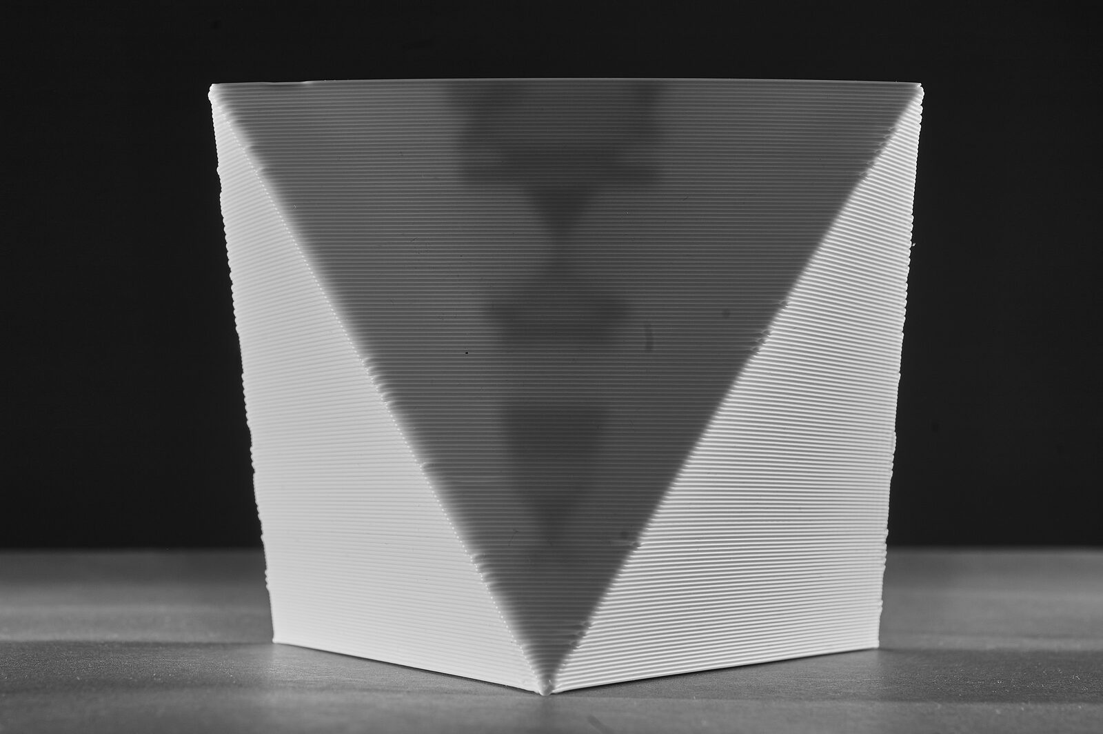 An octahedron resting on one face, lit from the sides, a faint V-shaped shadow of its internal structure rising through the wall