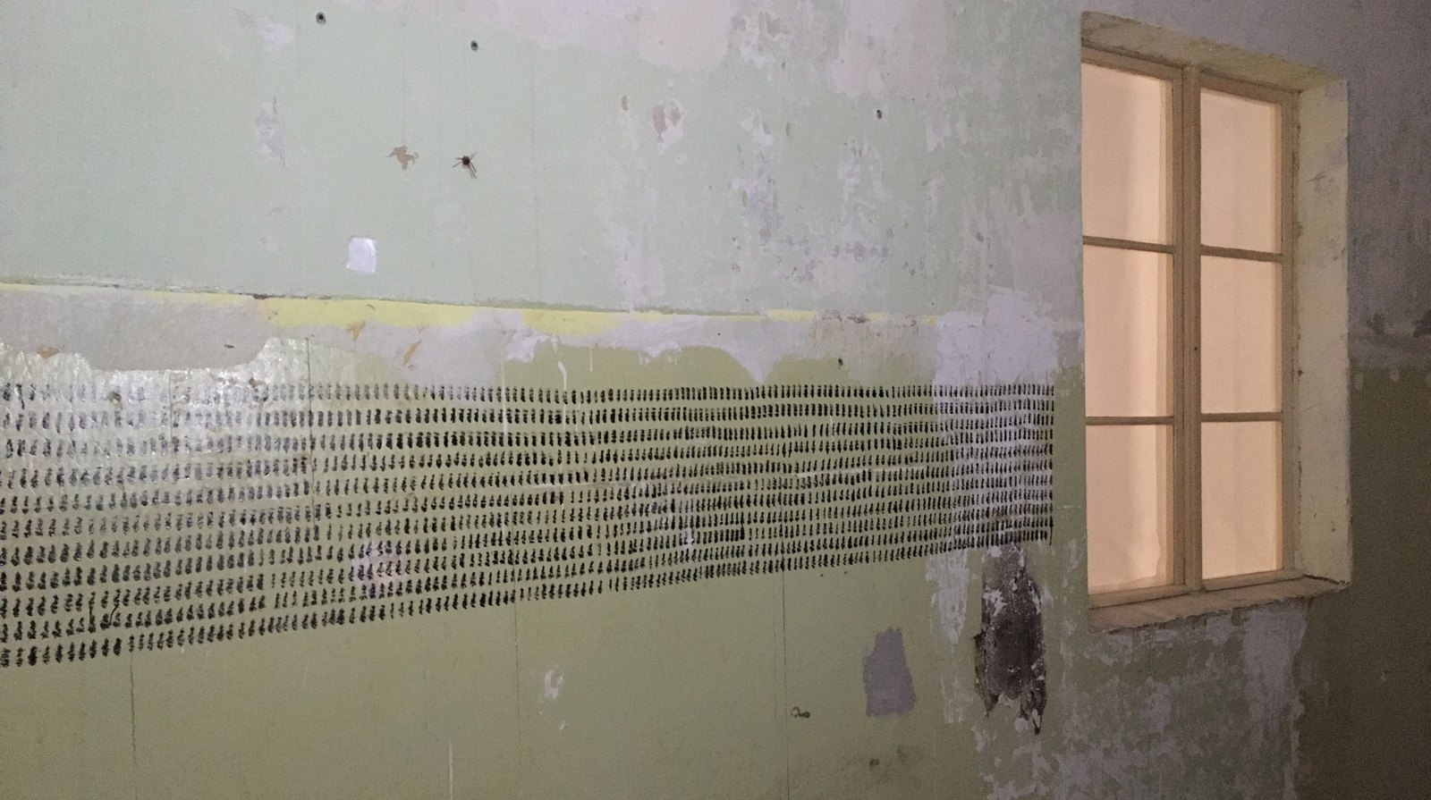 A peeling pale-green wall beside a curtained window, a broad band of short black brush strokes painted across it in close rows