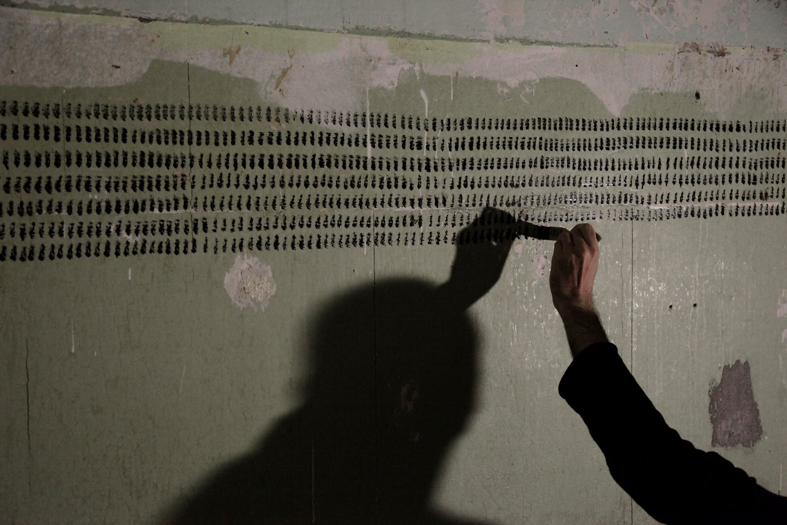 A hand pressing a brush to the wall at the end of a row of strokes, the artist's shadow thrown large across the rows