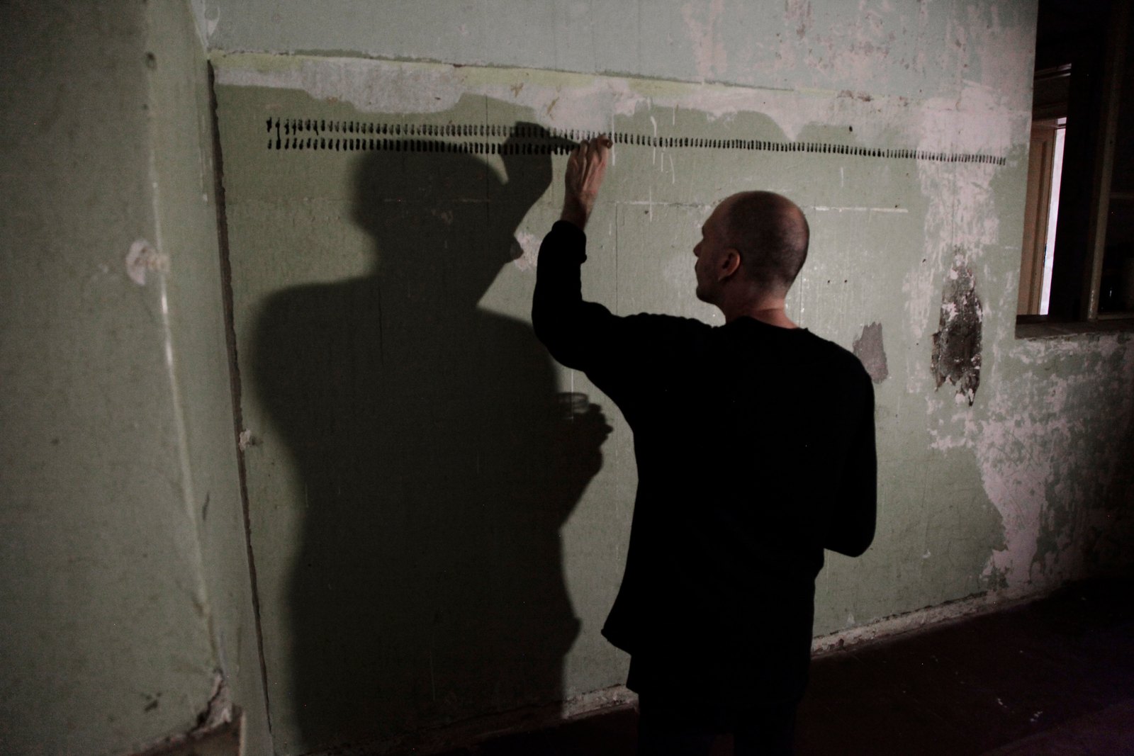 The artist in a dark room reaching up to the first row of strokes on a peeling wall, his shadow standing beside him