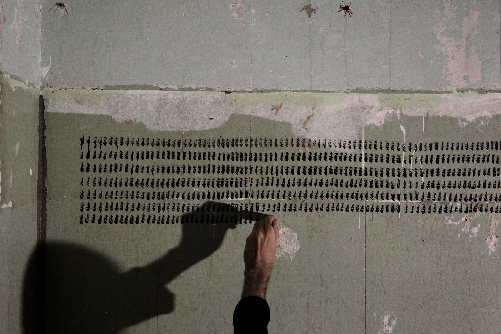 Rows of short black strokes on the bare wall, a hand under the lowest row and its shadow reaching along it