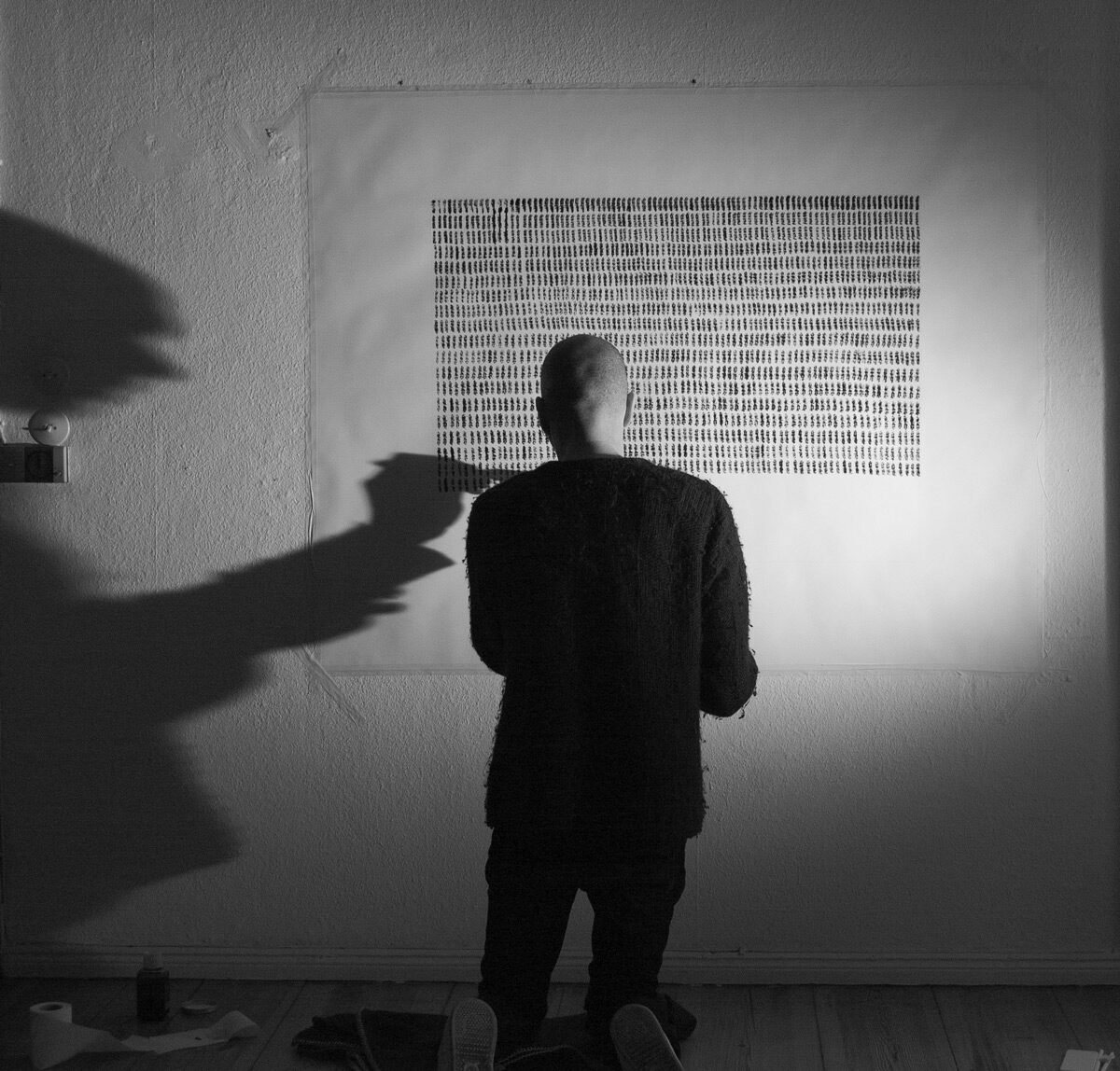 A figure standing before a large field of ruled strokes on a studio wall, his shadow thrown across it