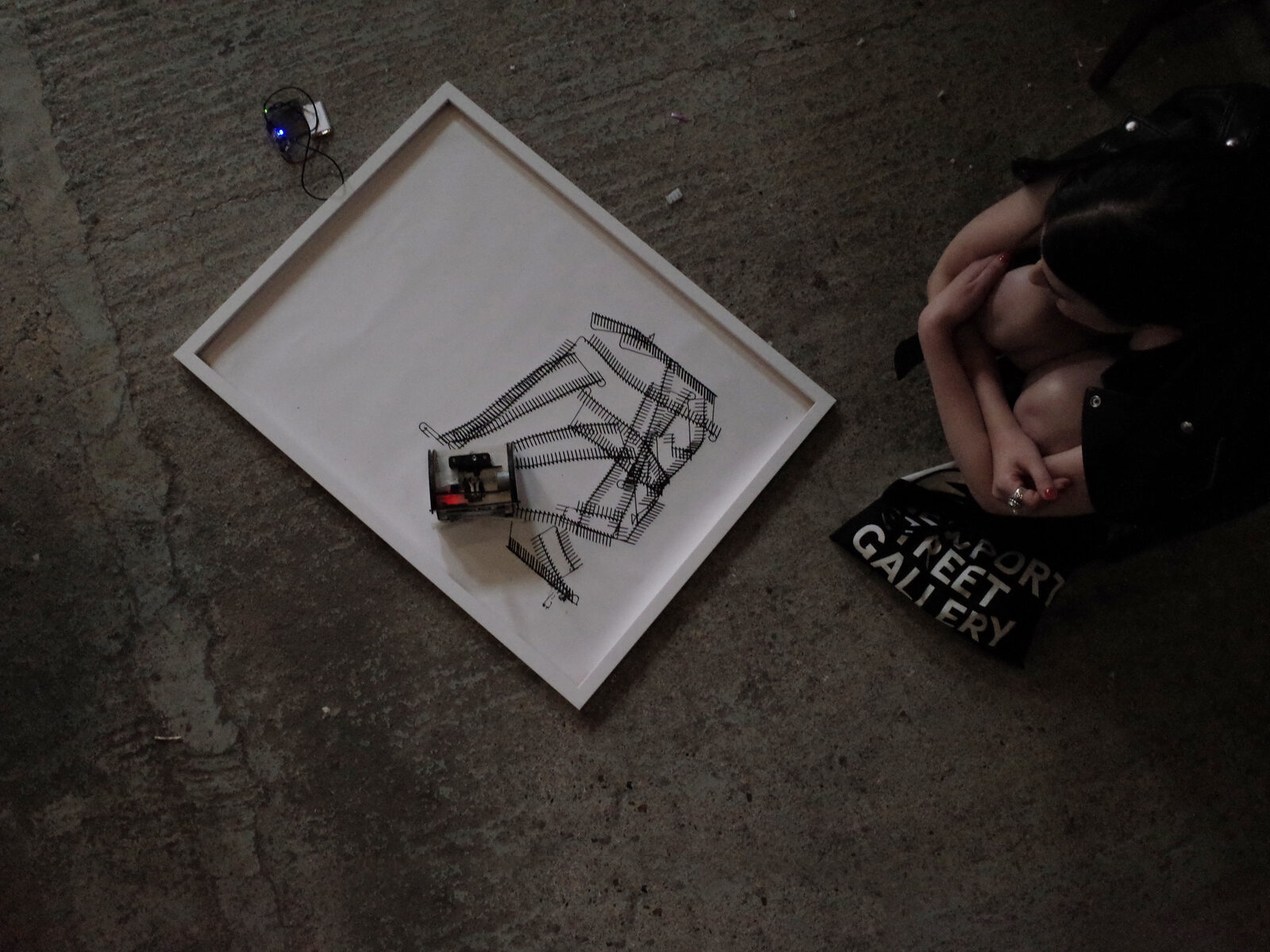 Seen from above: a framed sheet on a concrete floor with the small wheeled robot at work on it, a visitor crouching alongside