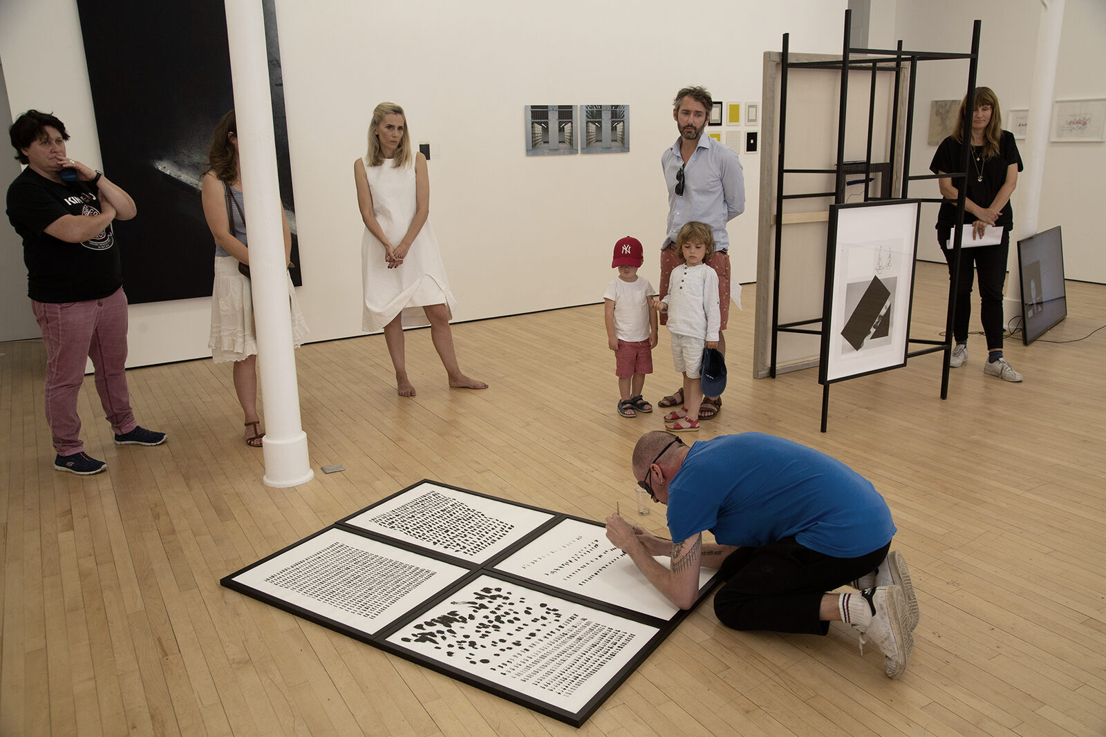 A gallery room with visitors and children watching the artist kneeling over framed sheets laid on the floor