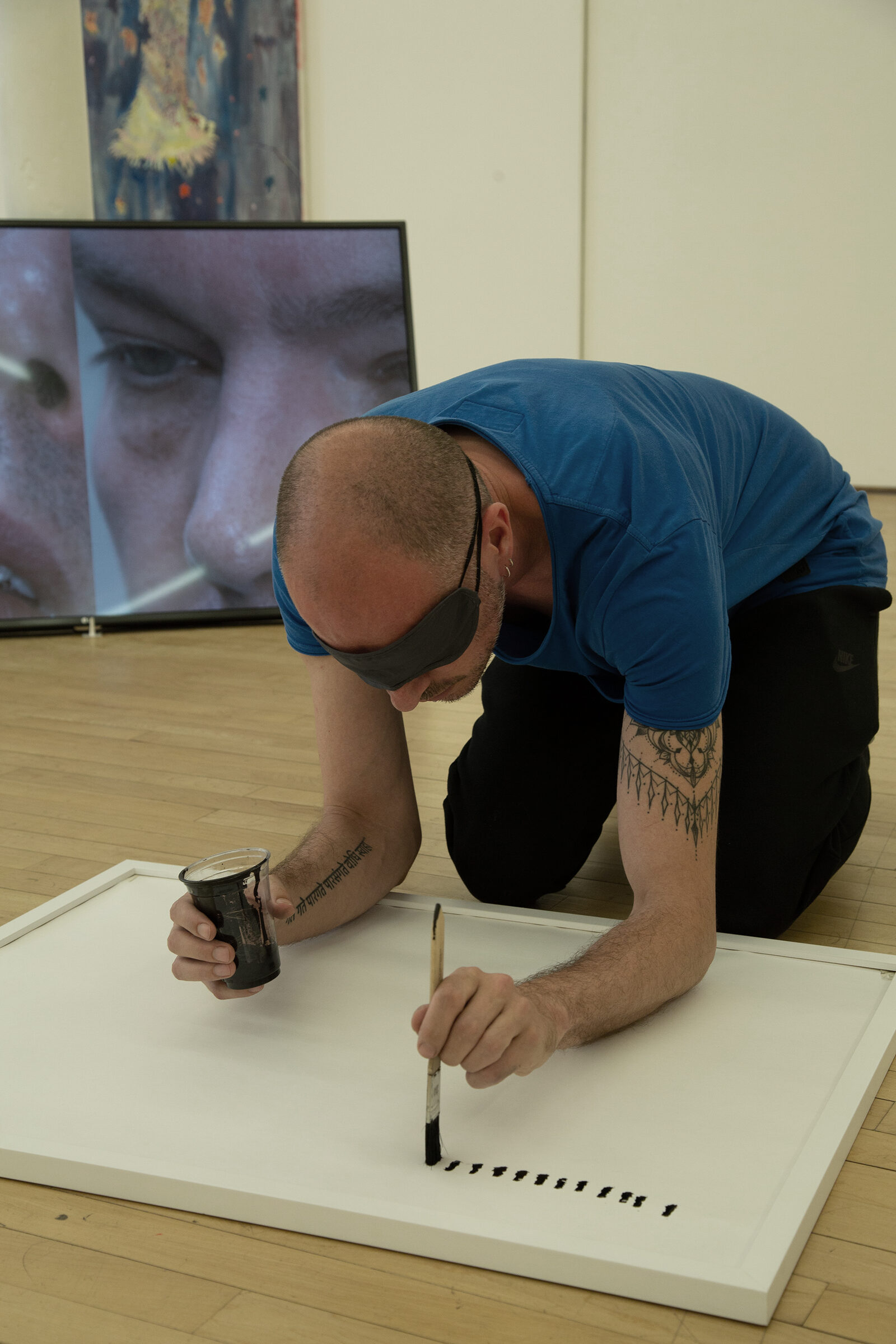 The artist kneeling close over a sheet, drawing by hand, a screen showing an eye behind him