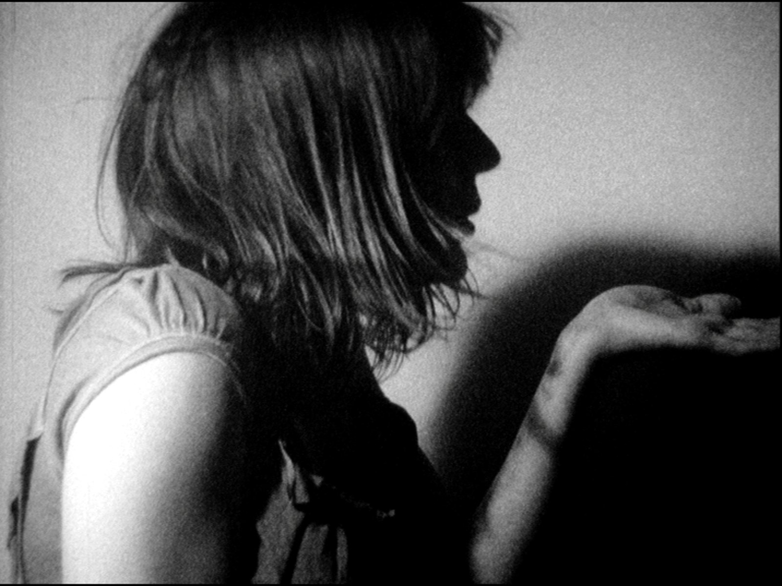 A woman seen from behind in grainy black and white, wet hair falling across her face, a puffed sleeve at the shoulder, one hand raised open into the light