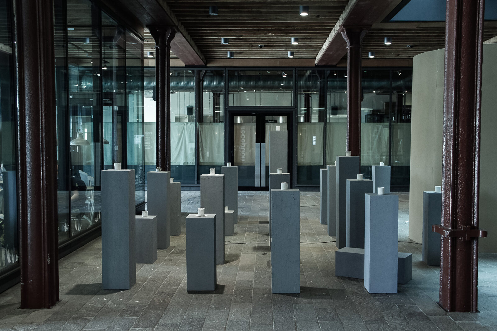 A large hall with rows of grey plinths, each carrying one small white cup, glass walls beyond