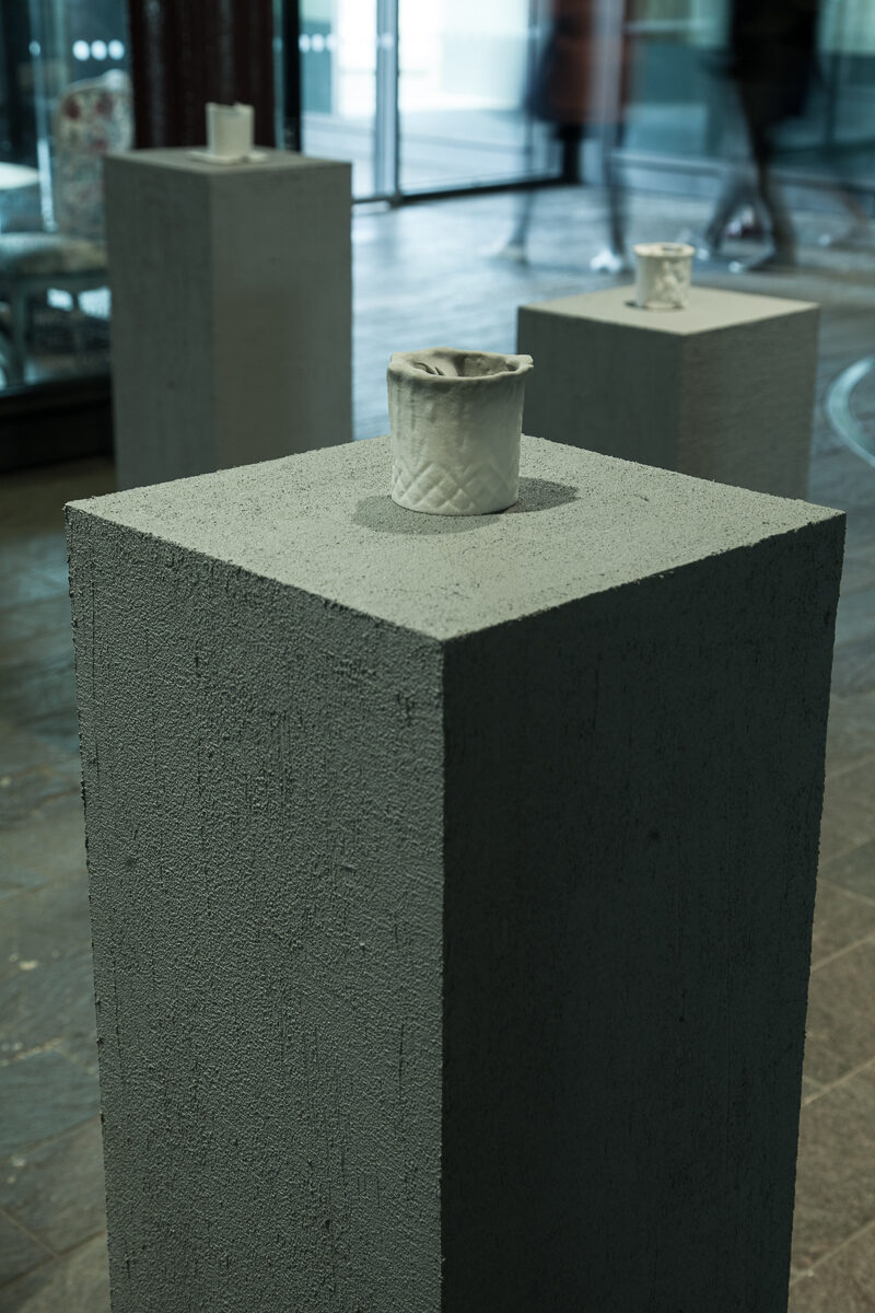 A single cup on a grey plinth with a second plinth behind it and figures passing beyond the glass