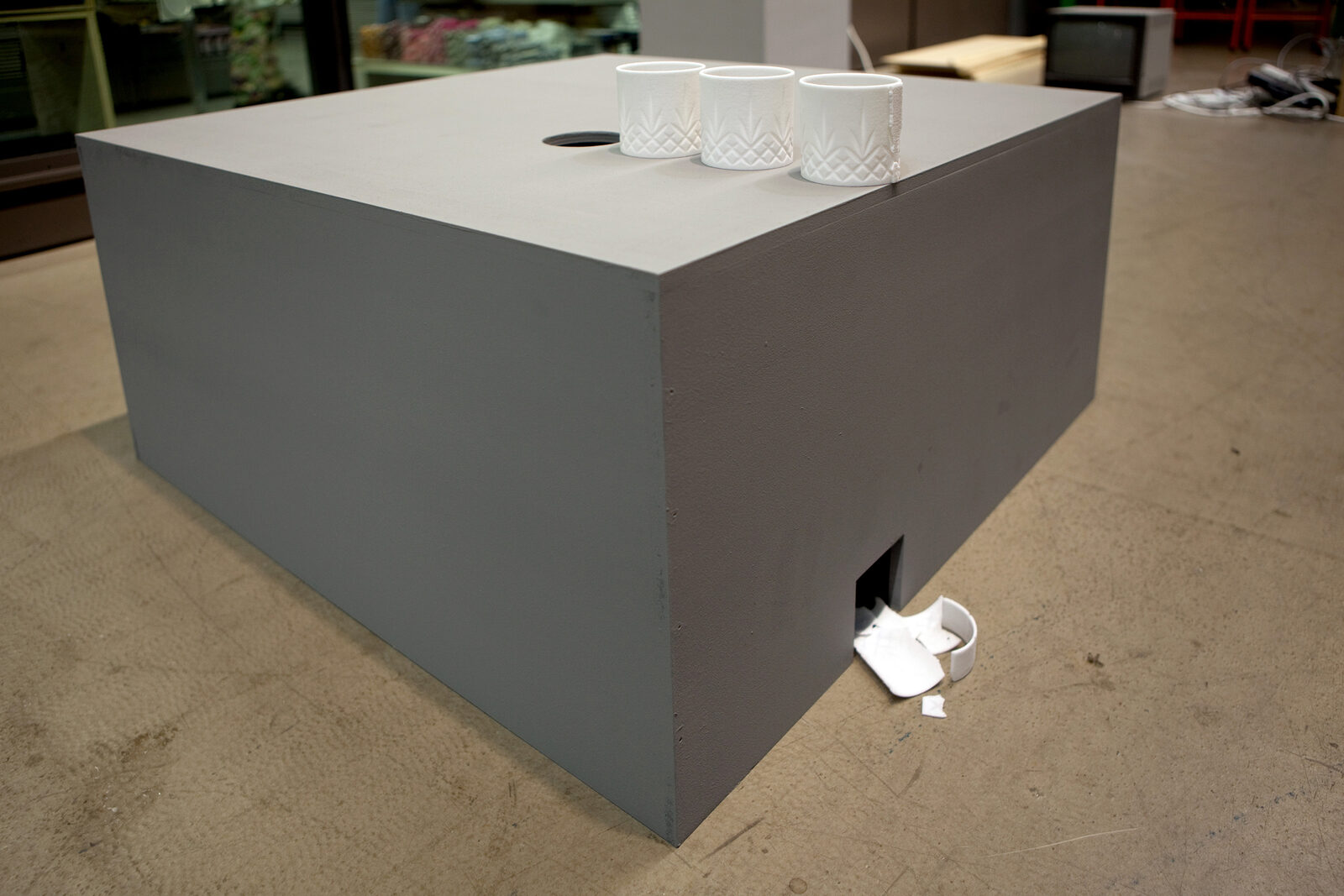 A large grey box on a floor with three cups on its top surface and one lying on the ground
