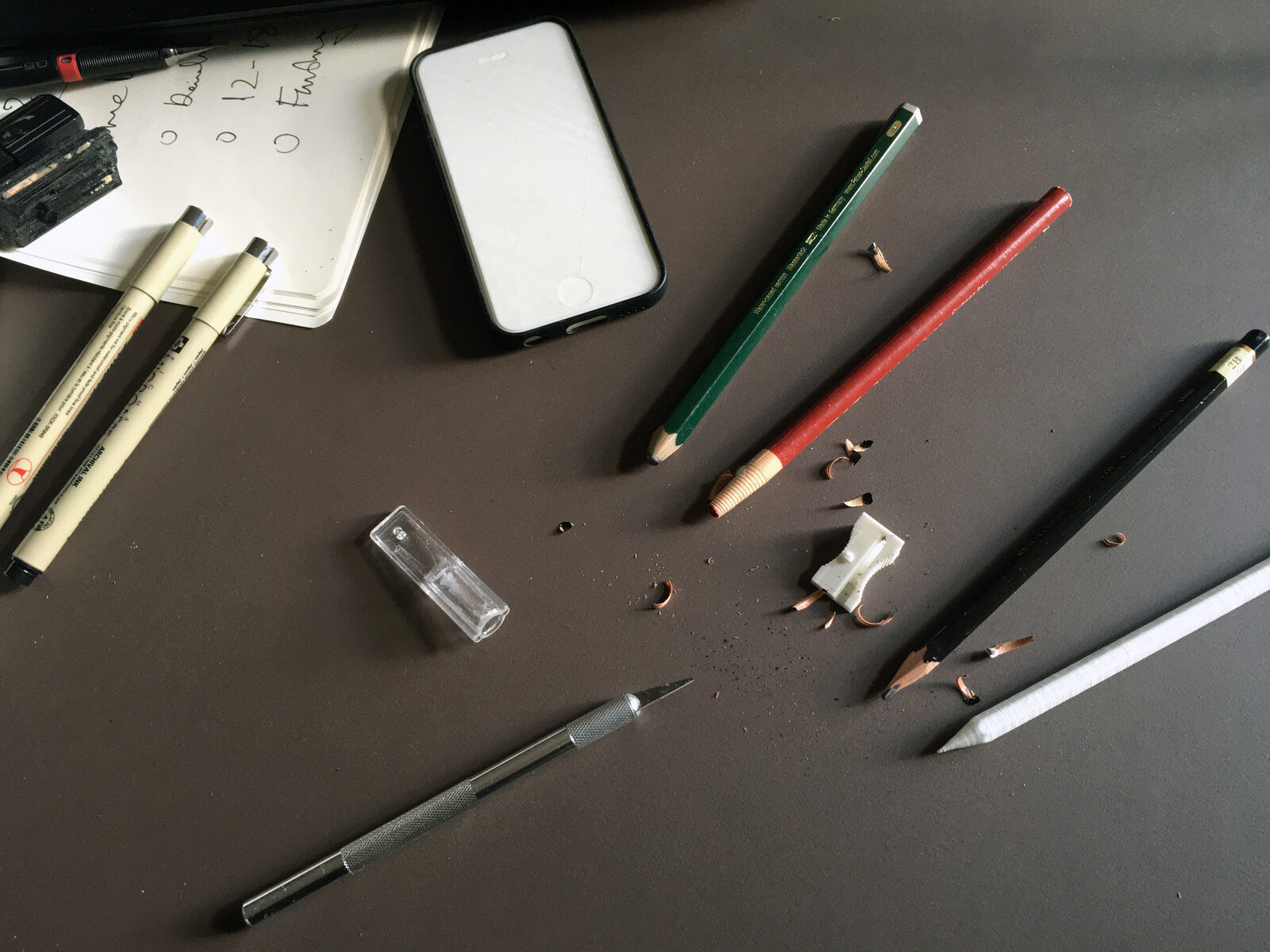 A desk with pens, pencils, a scalpel and shavings, among them a white printed pencil and a white printed sharpener