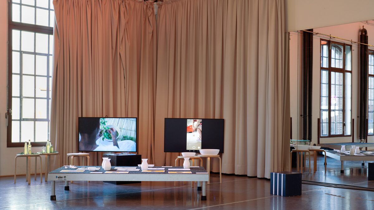 A long low table in a curtained hall, white 3D-printed vases and bowls among printed cards, two screens behind it and a mirrored wall reflecting the windows