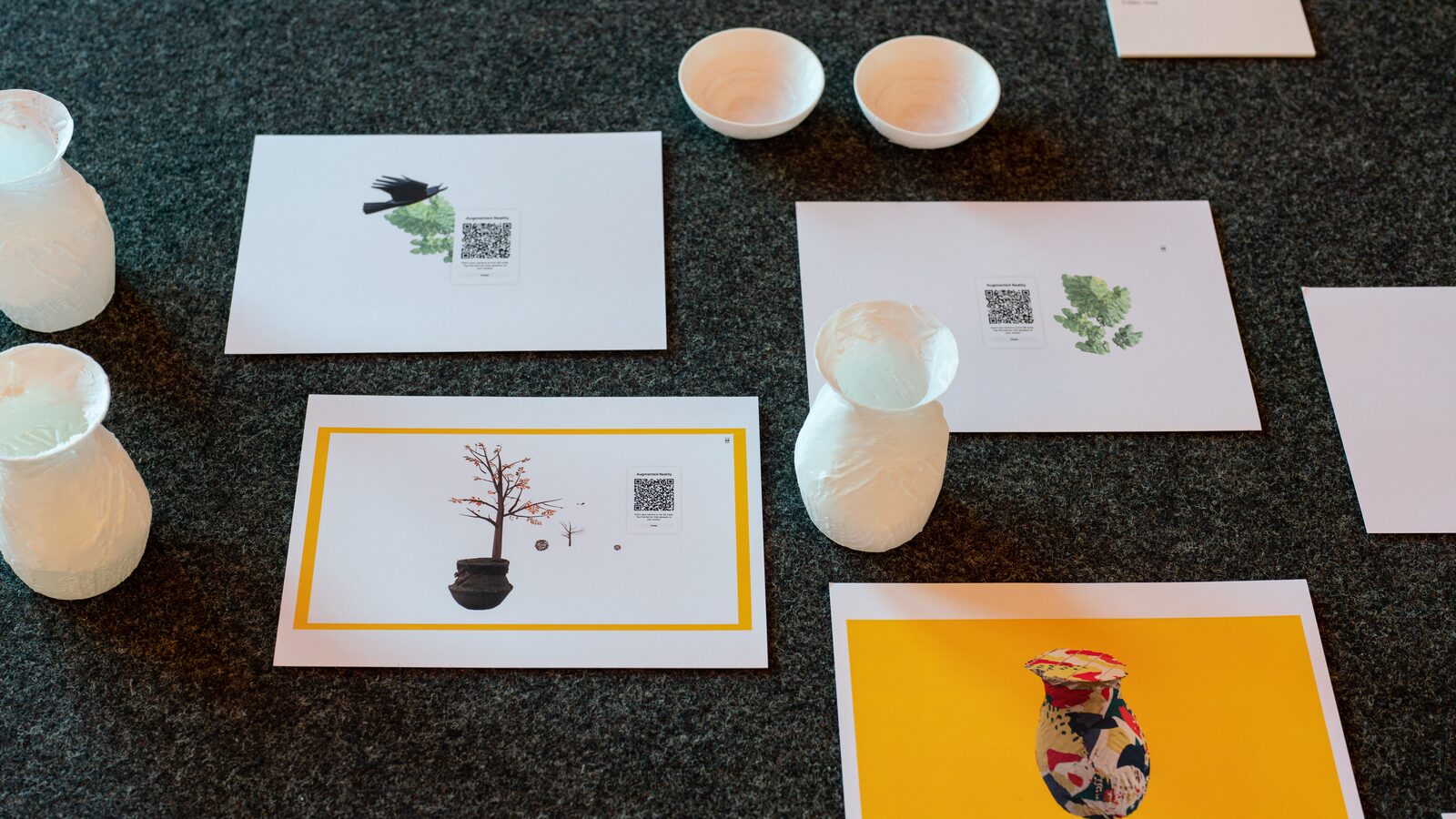 Seen from above on dark carpet: white 3D-printed vases and bowls beside printed cards with QR codes, a bare tree in a pot, a bird among leaves, a painted vase on yellow