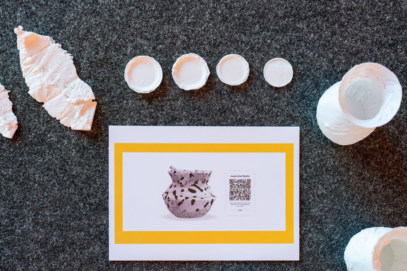 Seen from above on dark carpet: two white 3D-printed leaves, four small white bowls in a row, a vase on its side and a card with an augmented-reality QR code