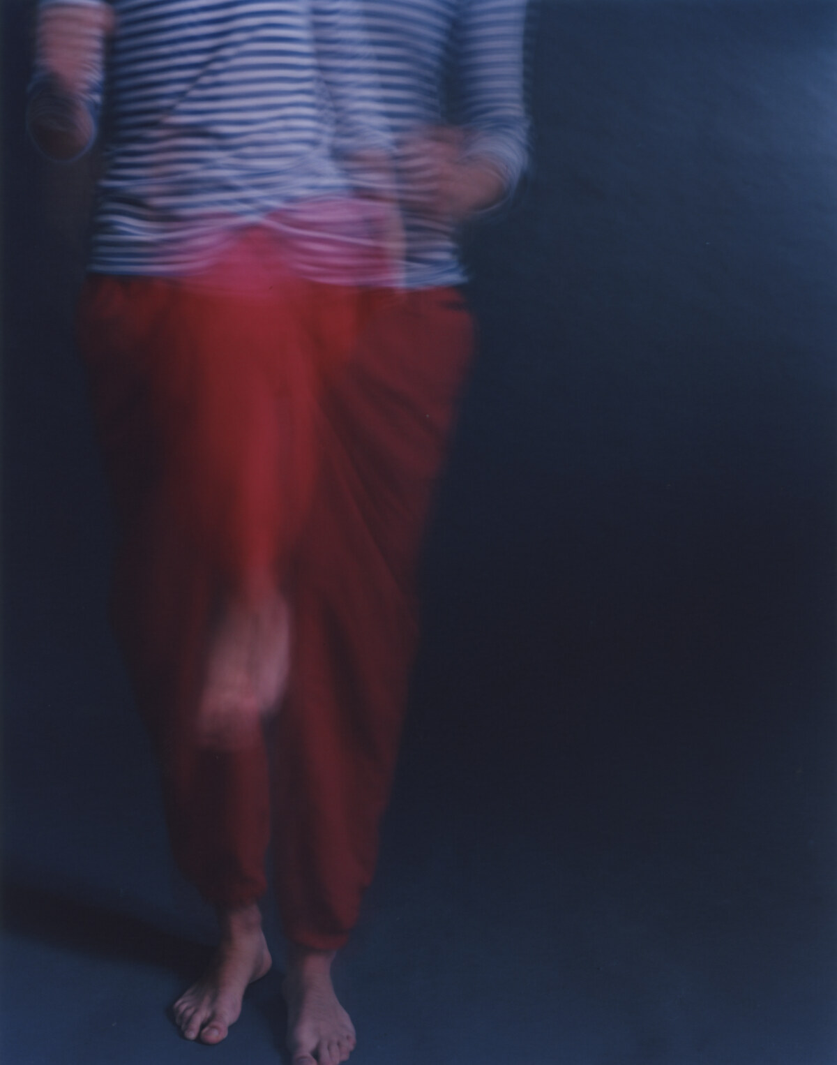 A standing figure, striped shirt above and red trousers below, both dissolved into vertical smears, bare feet resolving on the floor