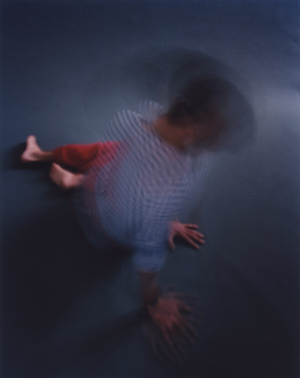The figure seen from above, crouched, both hands spread flat on the floor and repeated by the exposure, the head a dark mass at the top