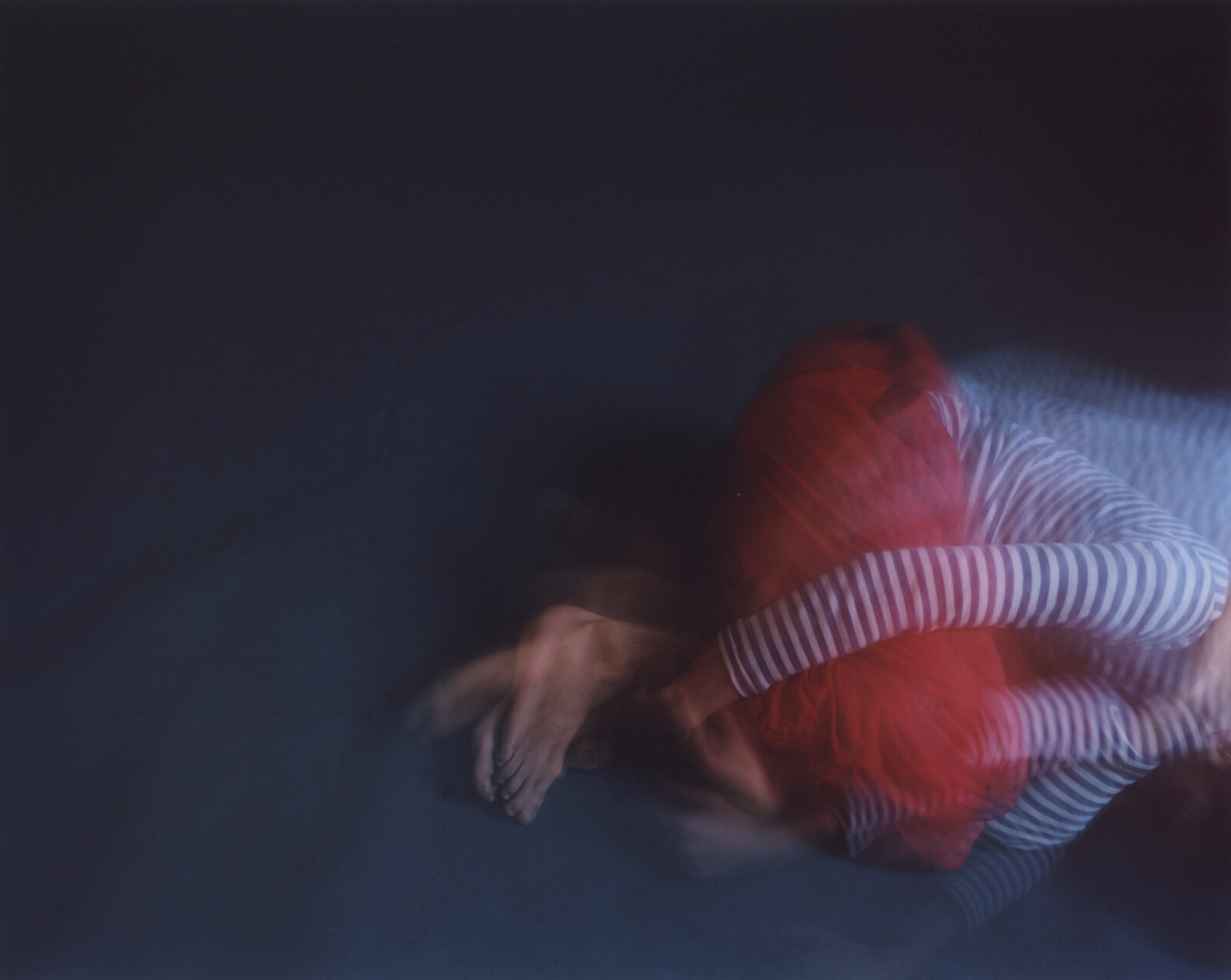 The figure lying along the floor, red and striped fabric run together into one mass, an arm extended away to the left