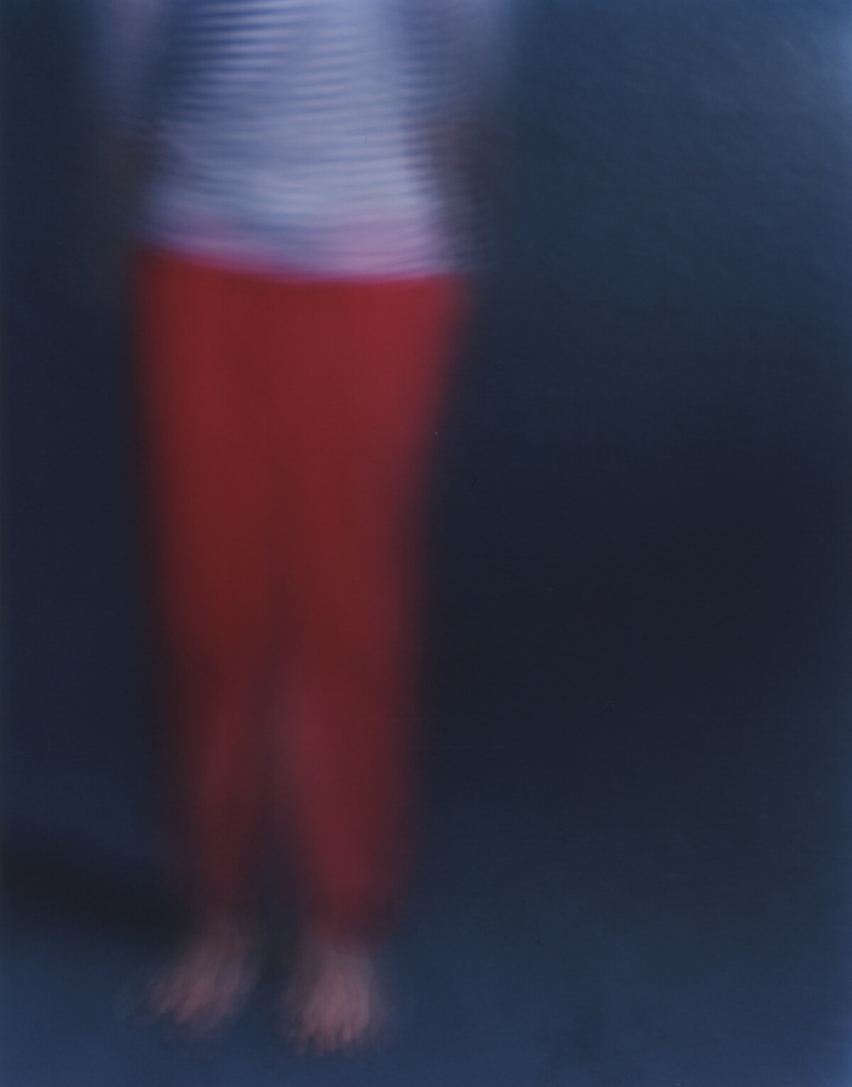 The figure upright and nearly still, red trousers holding their shape, the striped shirt vibrating above them, bare feet on the dark floor