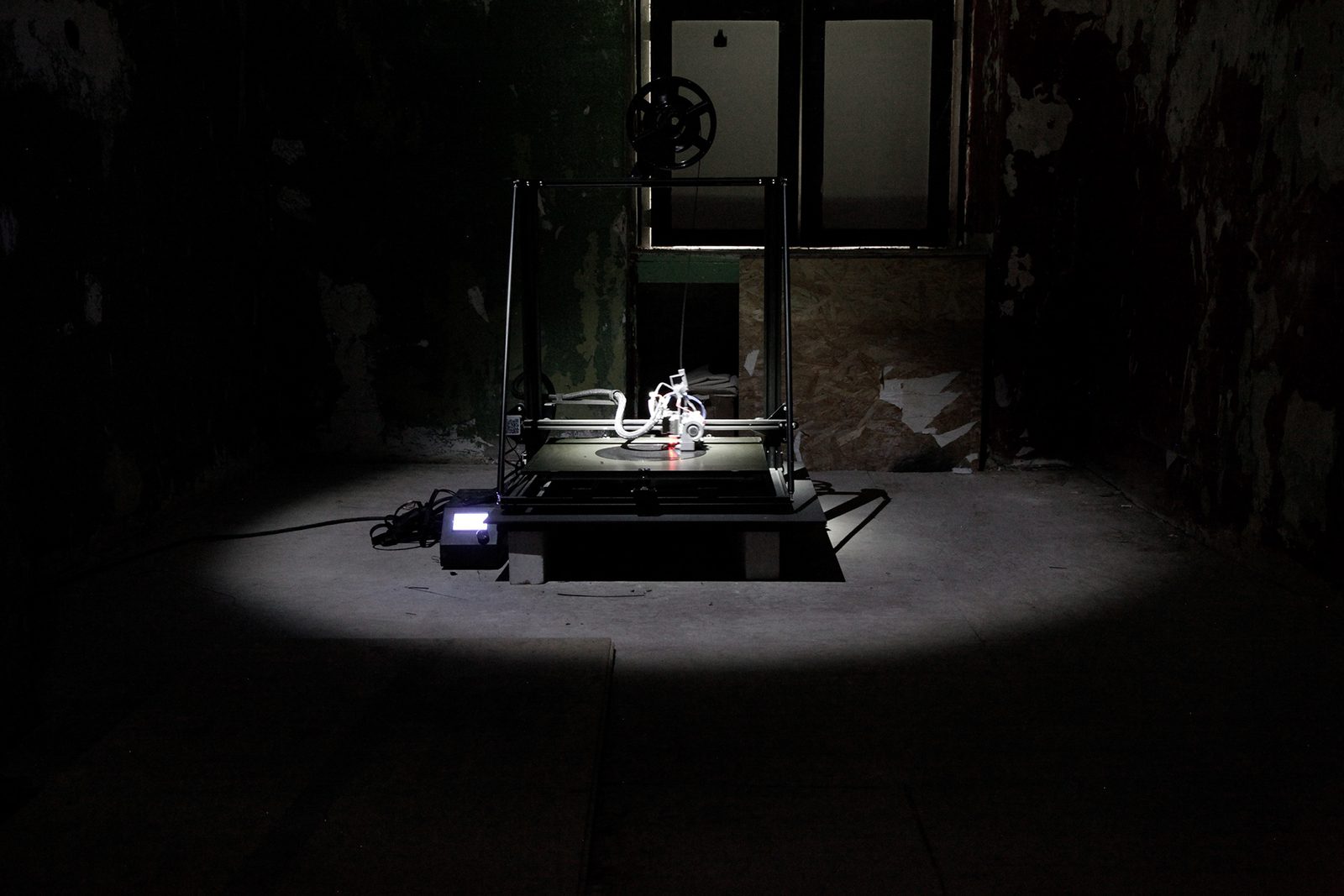 A 3D printer under a work light in a dark room with peeling walls, the floor around it still empty