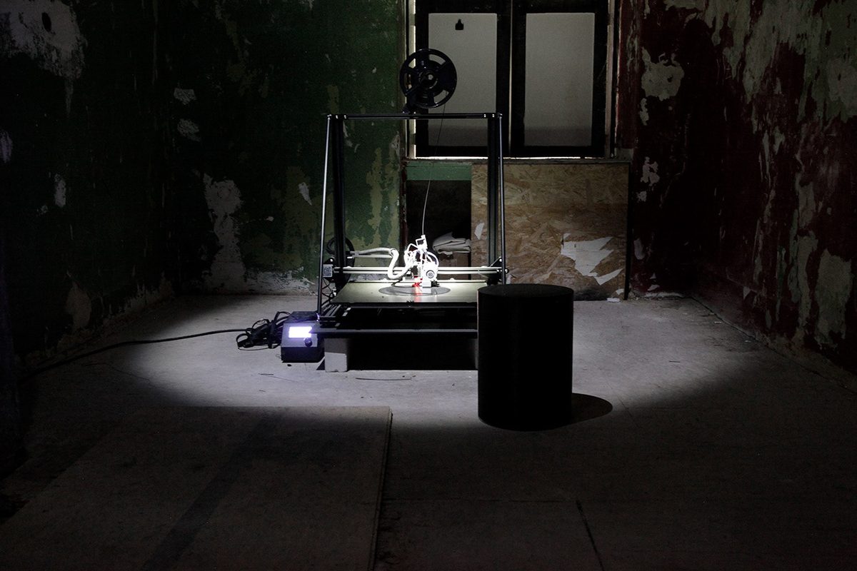 A 3D printer under a work light in a dark room with peeling walls, one printed seat standing at the edge of the light