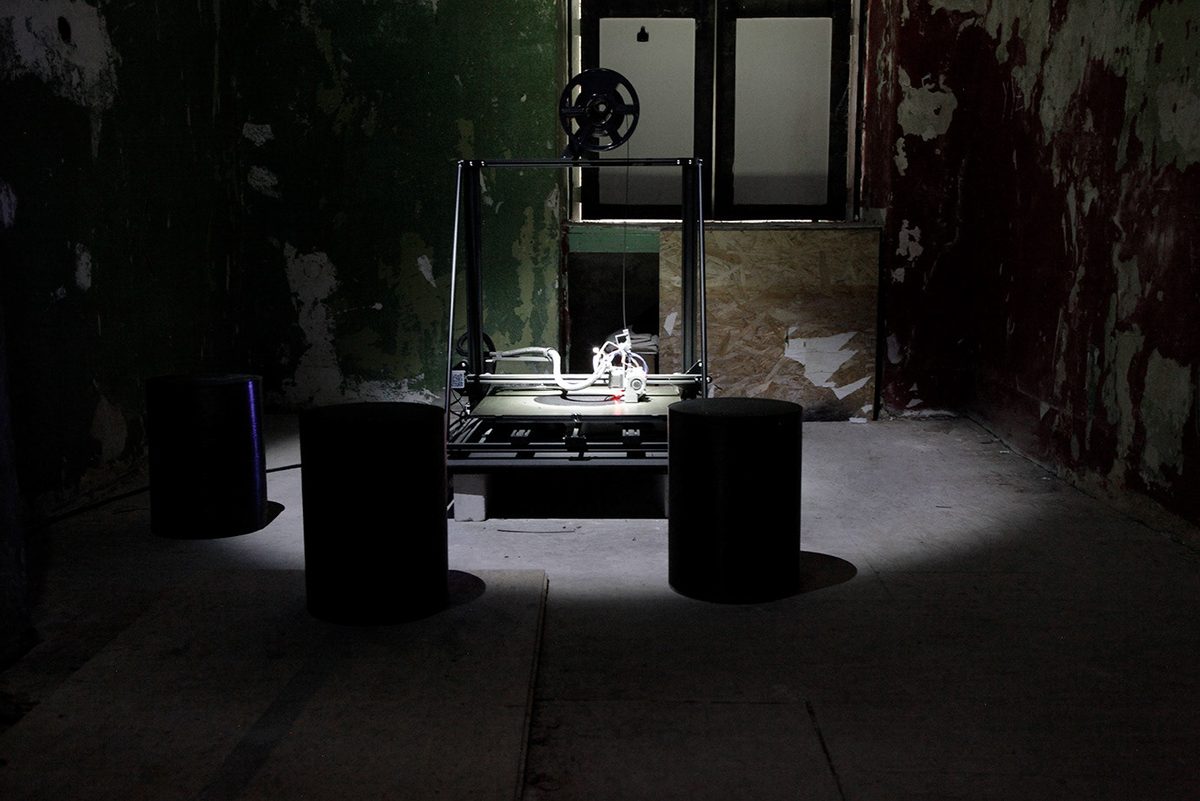 A 3D printer under a work light in a dark room with peeling walls, three printed seats around the printer