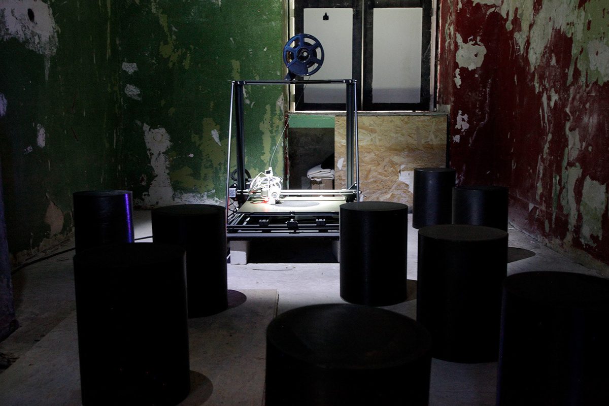 A 3D printer under a work light in a dark room with peeling walls, six printed seats filling the foreground