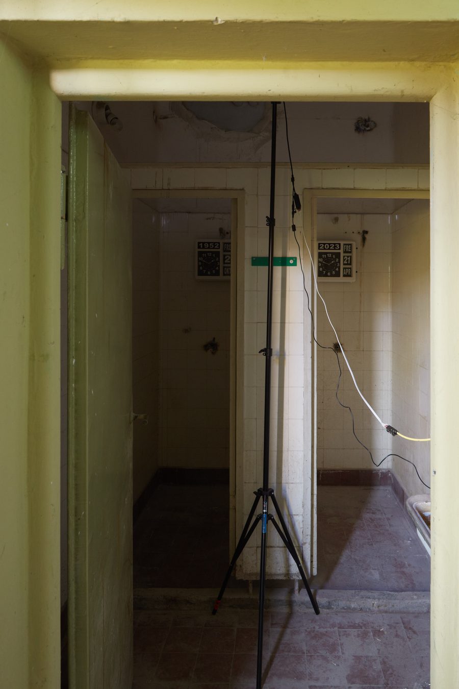 Two former shower cells seen through a doorway, white-tiled, a flip-clock on the back wall of each, 1952 on the left and 2023 on the right, a light stand and cables in the passage between them; the rooms closed to visitors