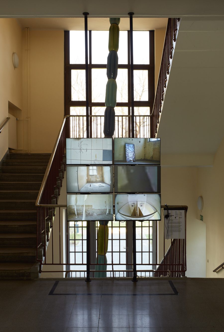 The six-screen wall in the foyer seen from the front against the stairwell window: six monitors on two black poles, each showing the live feed of one basement room, the lower right one the two clocks