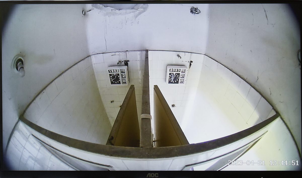 A surveillance monitor showing a fisheye view down into the two former shower cubicles, the 1952 clock in one and the 2023 clock in the other, a timestamp burned into the corner