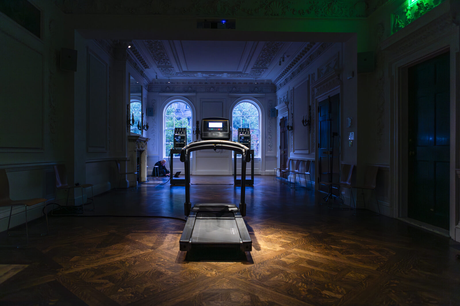 The full length of the panelled hall in blue light, the treadmill on the parquet in the foreground and the two printers standing between the arched windows at the far end