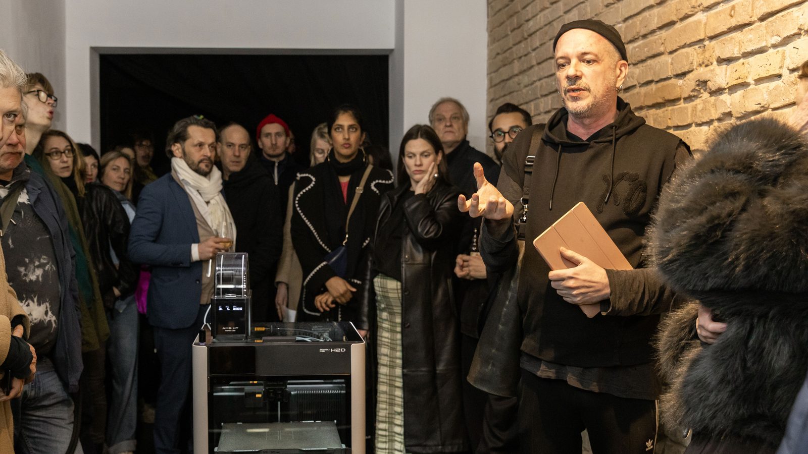 The packed gallery on the opening night: visitors pressed along the brick wall and into the doorway, a man in a black beanie speaking with a notebook in his hand, the printer among them