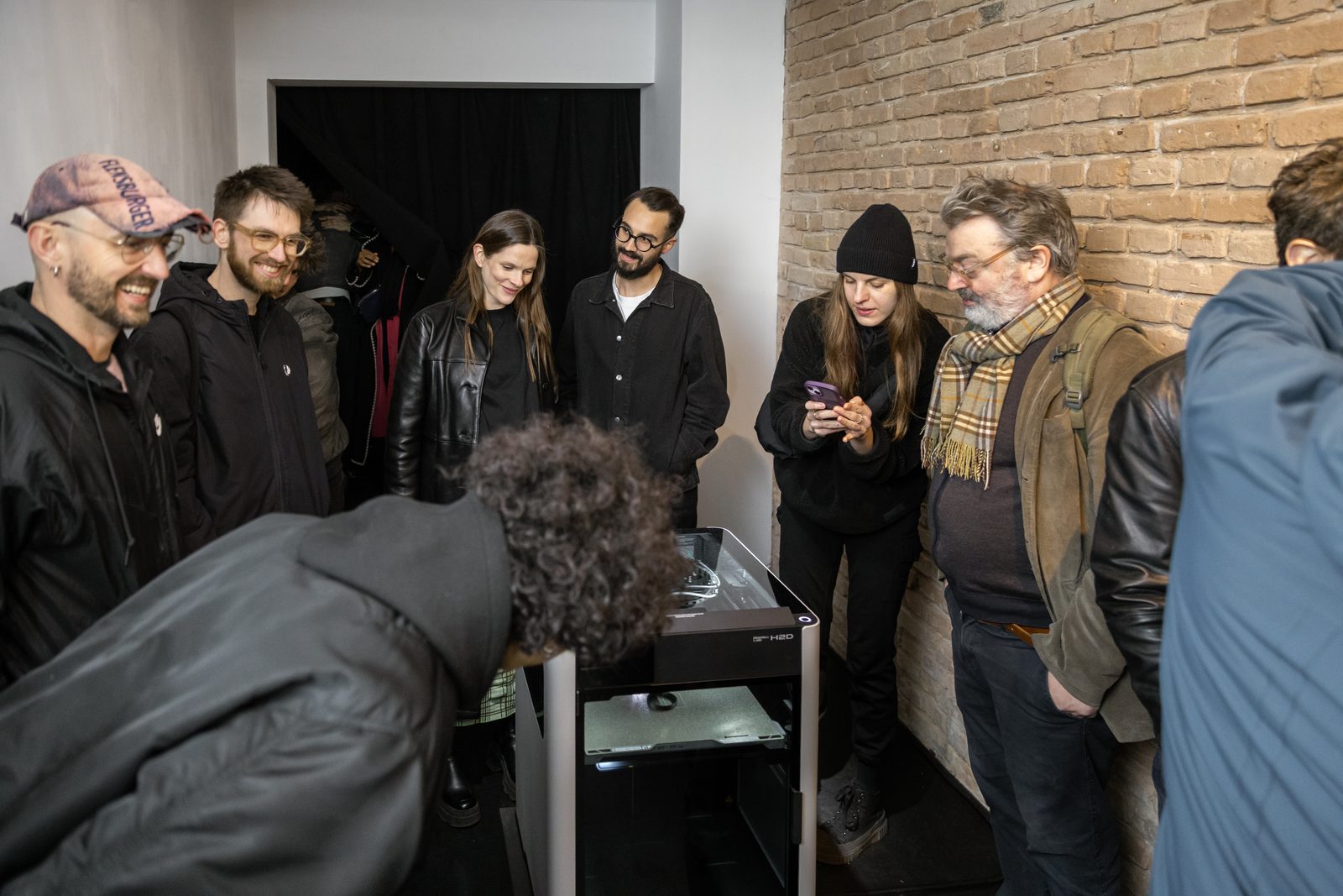 Visitors crowded round the printer by the brick wall, one bent over the machine to look inside