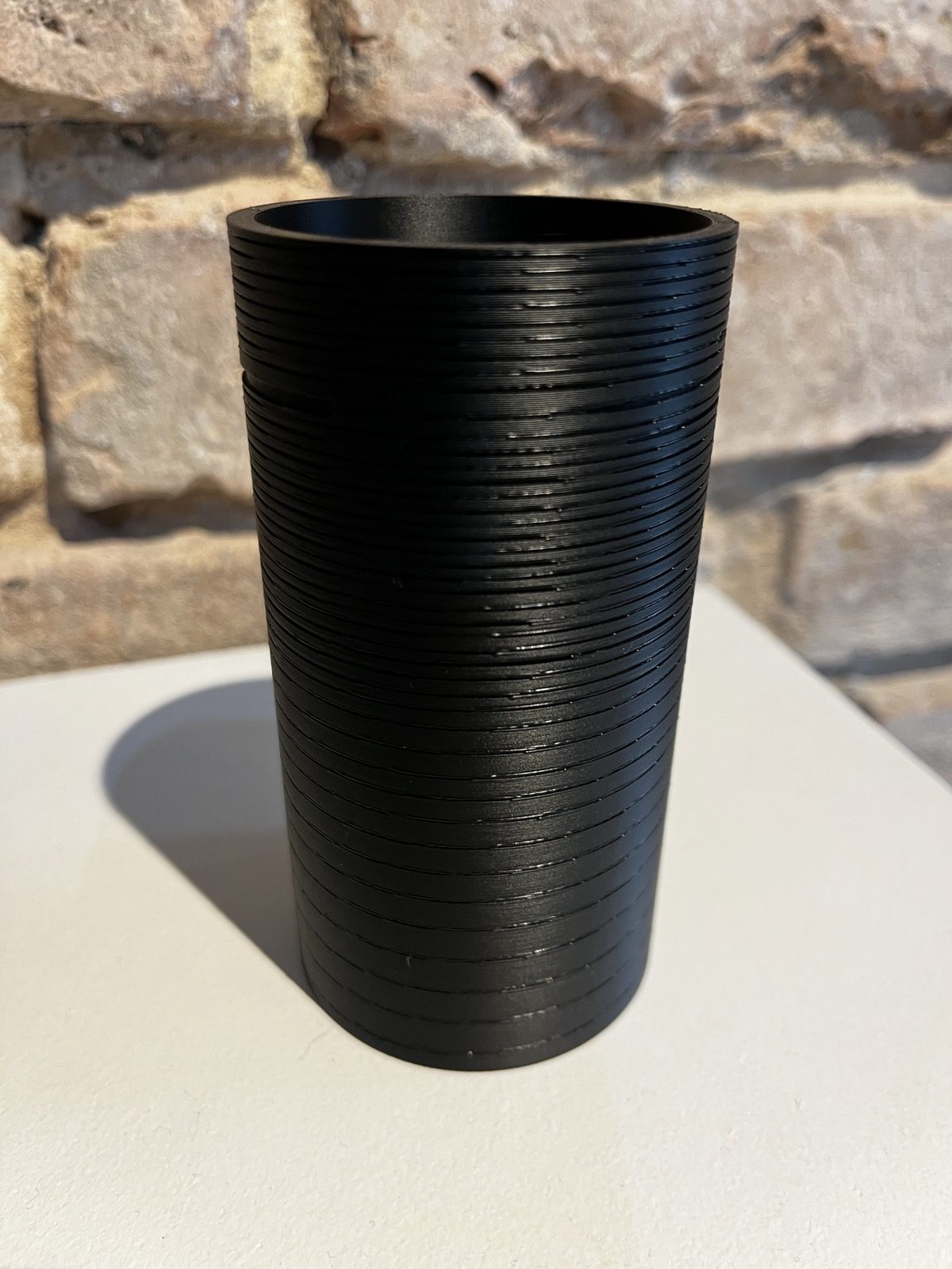The same cylinder printed in black on a white plinth against a brick wall, its spiral groove dense and nearly even from foot to rim