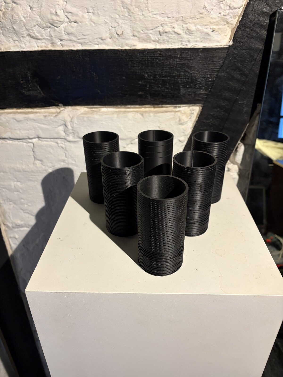 Six black cylinders on a white plinth against whitewashed brick and a black beam, each carrying a different surface: fine ribs, dotted bands, wide grooves, one nearly smooth