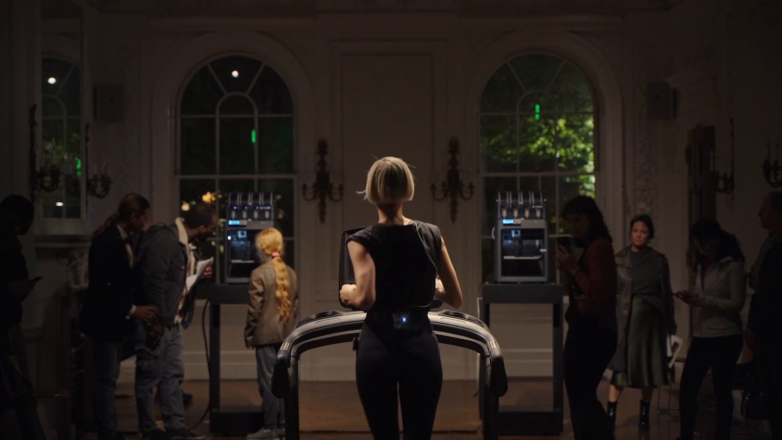 Katz running on the treadmill seen from behind, between two printers under the arched windows; on the left two men and a girl with a long braid turn to one printer, on the right visitors watch her