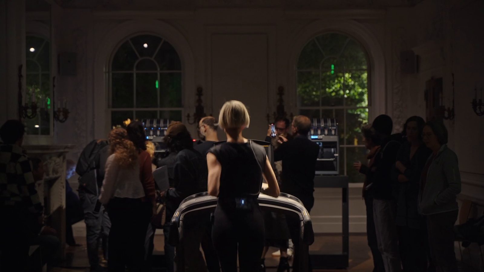 Katz running on the treadmill seen from behind while visitors crowd round both printers under the arched windows, some filming the chambers with their phones