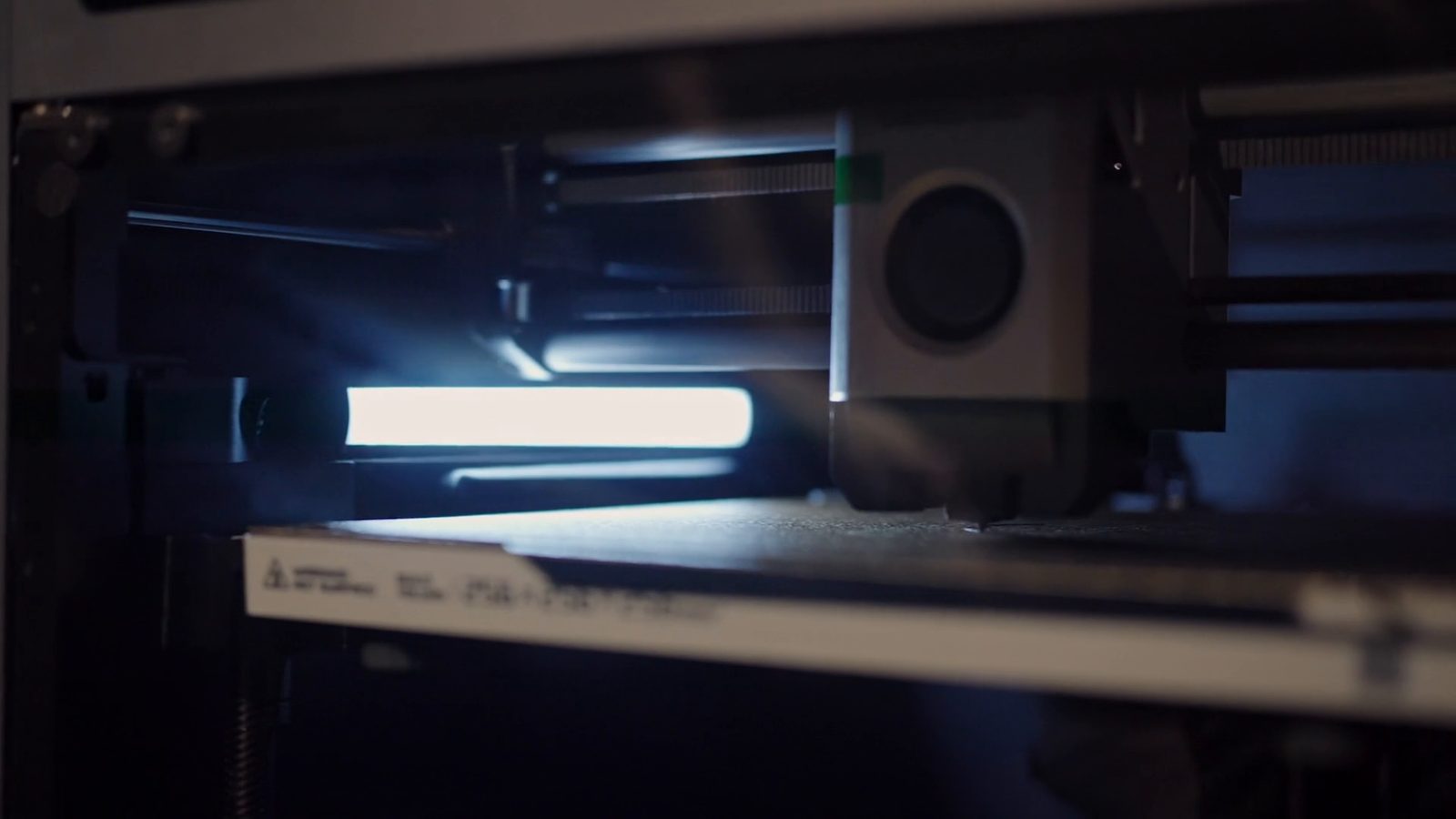 Inside the printer: the print head lowered to the textured plate as a print begins, lit from the left by a bar light