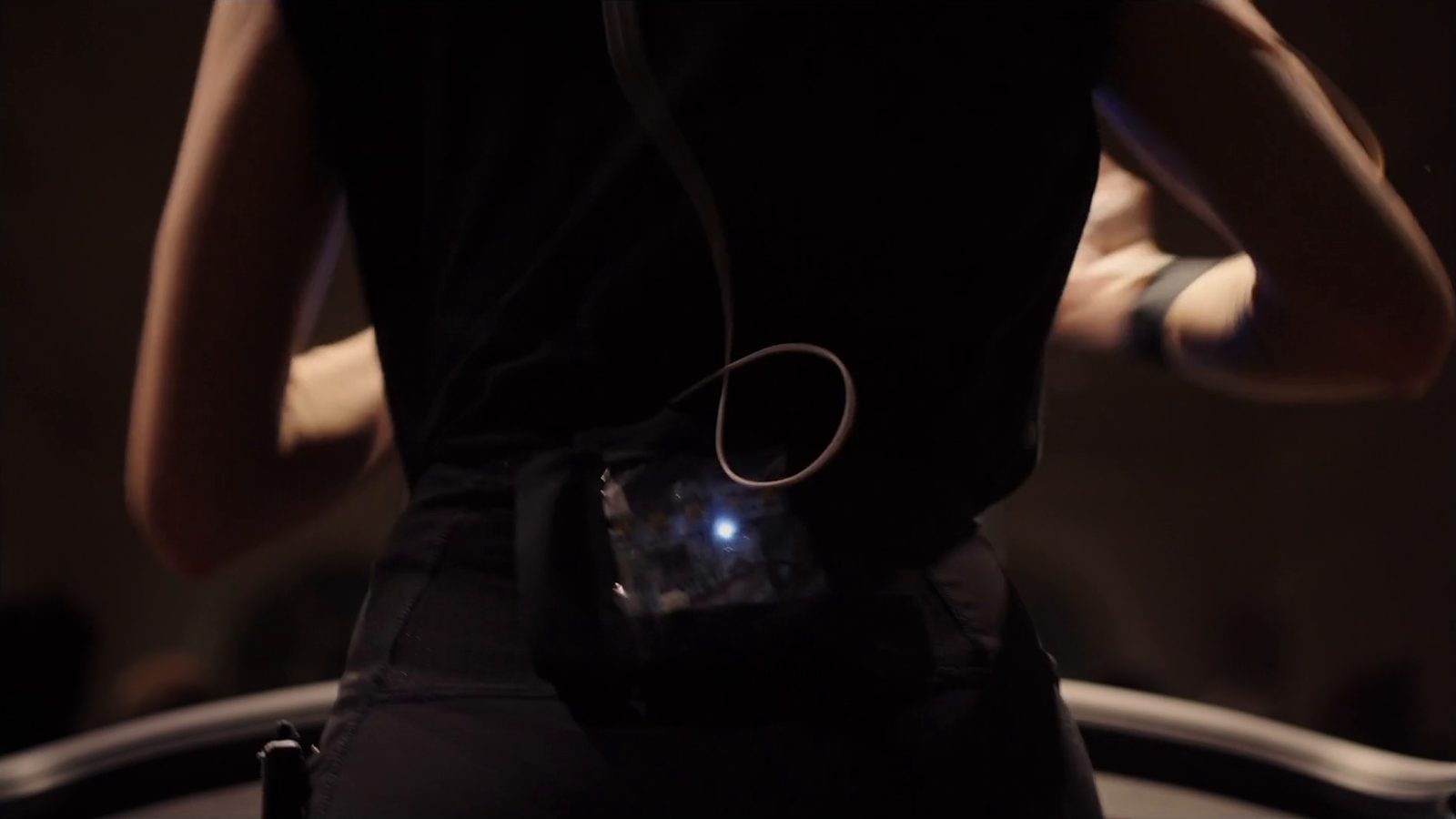 Katz seen from behind in a black top while she runs, the sensor box at her waist lit blue, its cable looping up her back, both arms bent in mid-swing