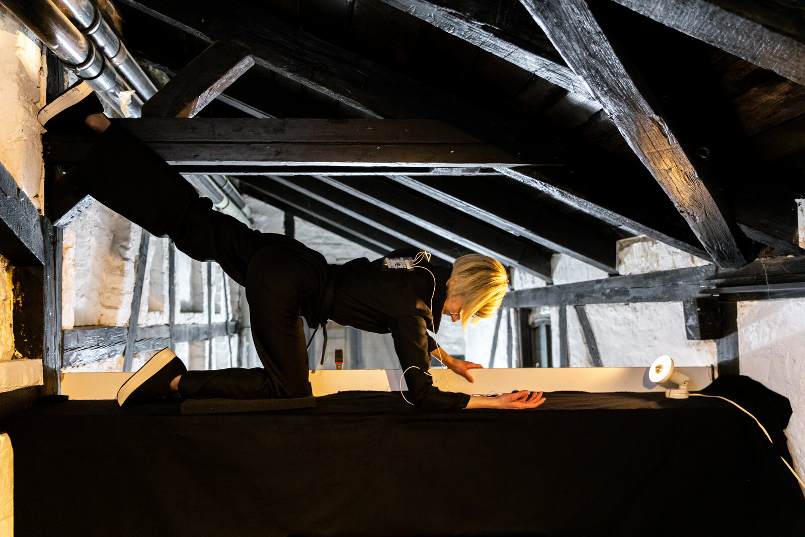 Katz stretched along the length of a roof beam under the dark timbers, one hand reaching to the far edge, the leads trailing from her